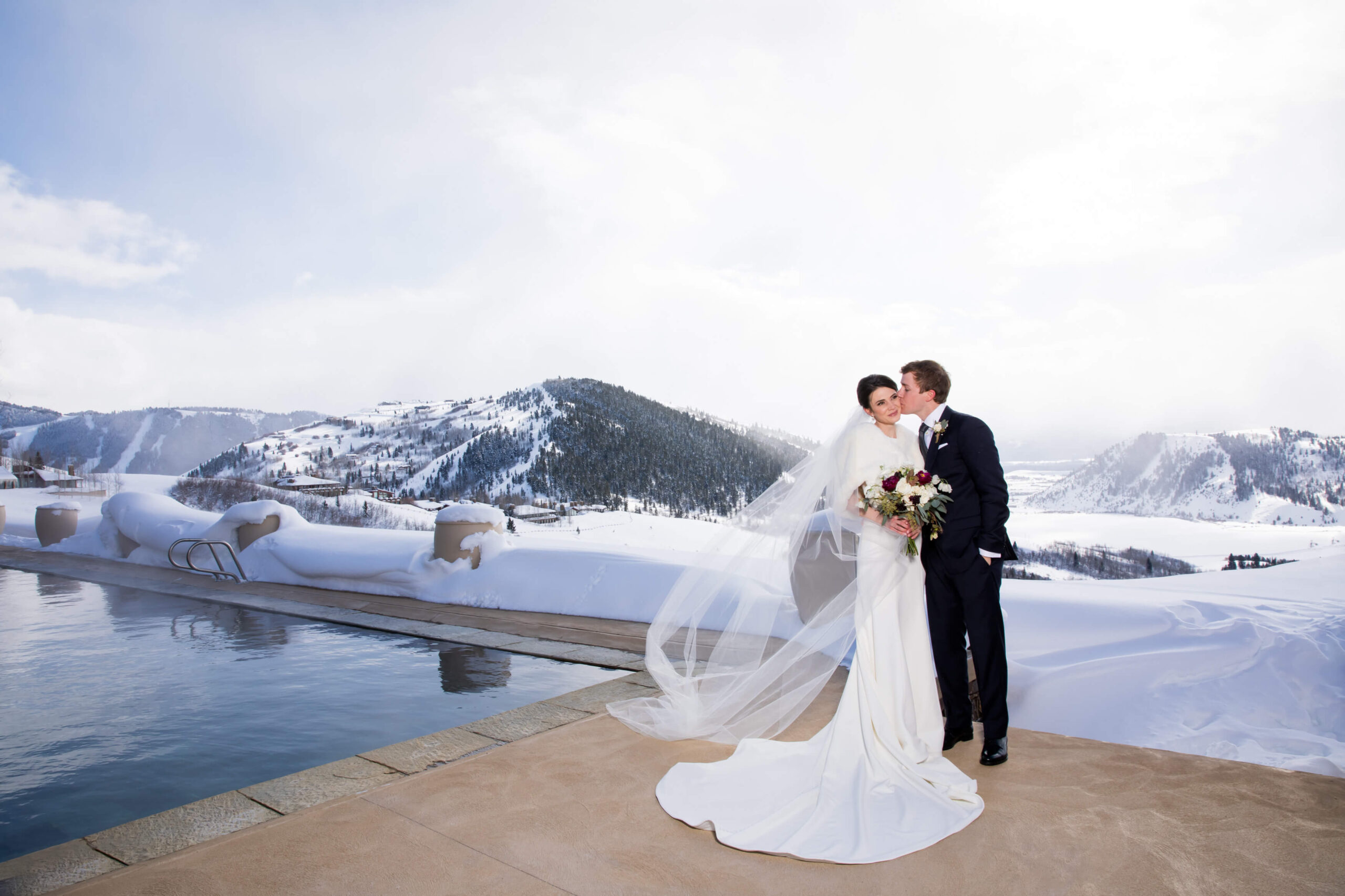 The bride and groom stand together by the pool at Amangani in Jackson Hole, framed by snow-covered mountains. The bride’s veil flows in the breeze as she holds a bouquet, highlighting their elegant winter wedding.